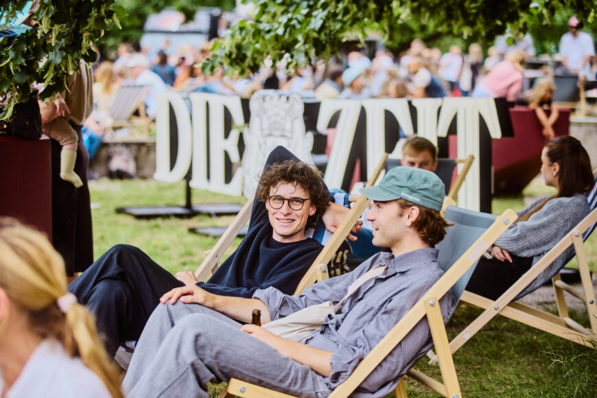 Zwei junge Männer lachen in Liegestühlen sitzend vor dem ZEIT-Logo auf dem Campus der Uni Hamburg.