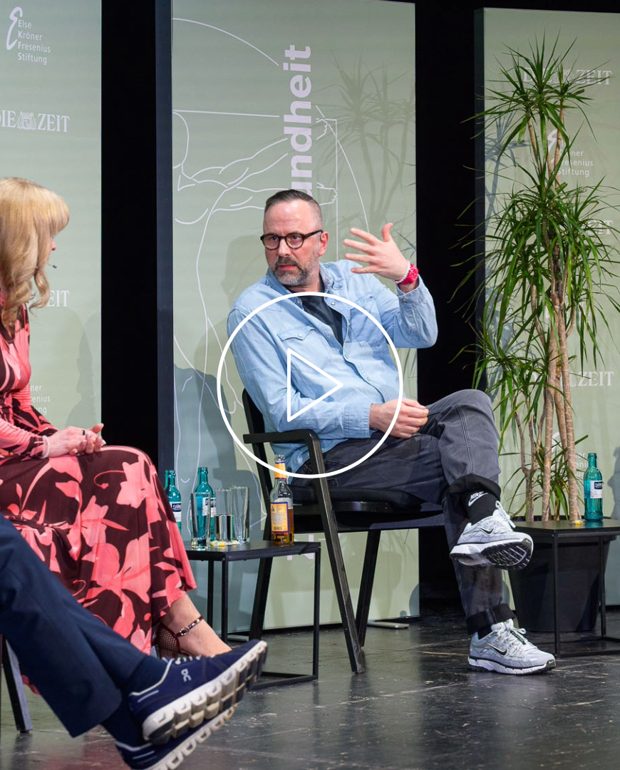 ein Podium mit vier Personen, von links nach rechts, Ulrich Hegerl, Eva-Lotta Brakemeier, Kurt Krömer und die Moderatorin Nora Burgard-Arp. Kurt Krömer spricht gerade und hebt gestikulierend den linken Arm. Das Podium sitzt vor pastelligen, olivfarbenen Aufstellern. Eine Pflanze ist auch zu sehen